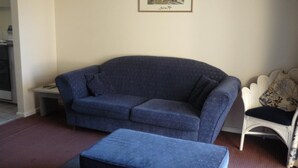 Una sala de estar con un sofá azul, un reposapiés a juego, una silla blanca con un cojín floral y un cuadro en la pared.