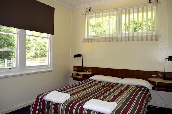 A hotel room with a bed, a nightstand, a window with blinds, and a view of greenery outside. - Blackheath Holiday Cabins (Blackheath)