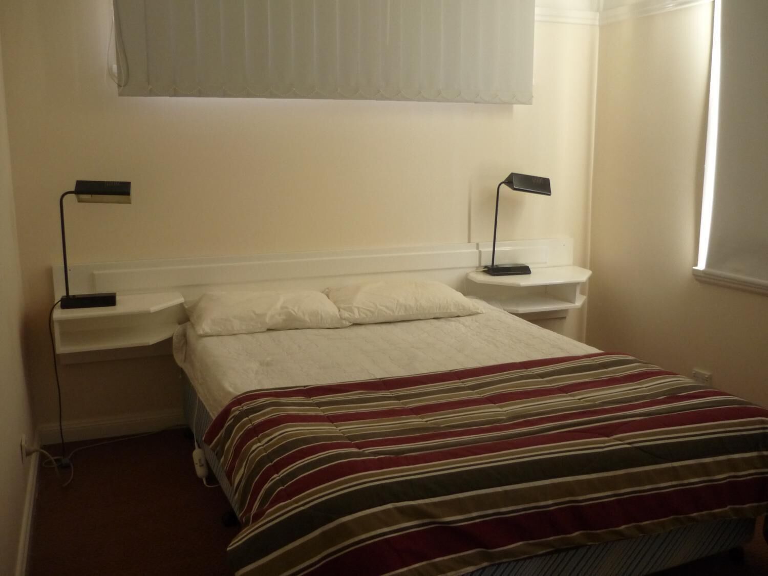 A simple, clean bedroom with a bed, two bedside tables, and a window with blinds.