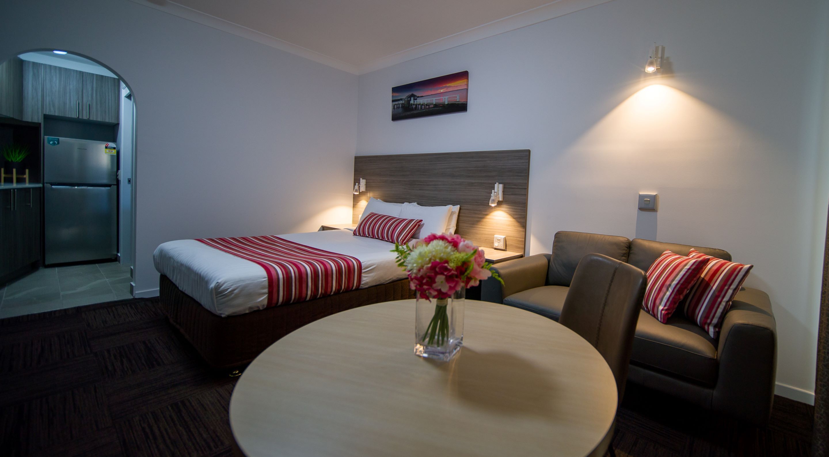 Une chambre d’hôtel comprenant un lit, un canapé, une table avec un vase, un tableau au mur et un coin cuisine.