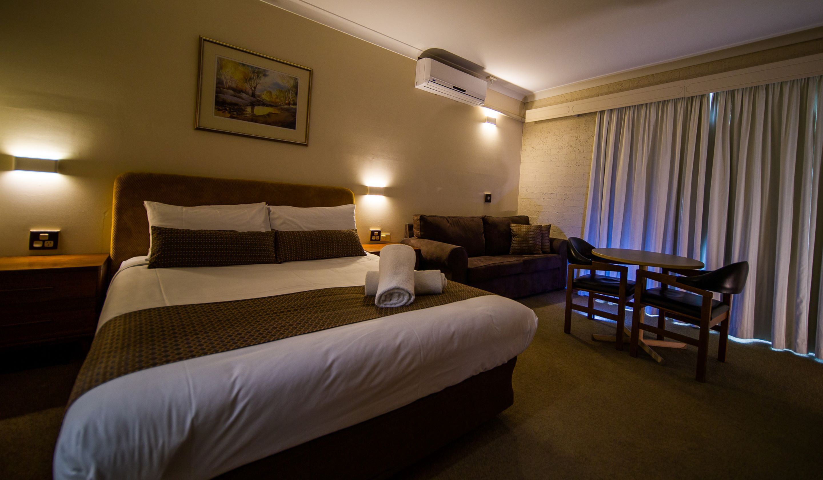 Habitación de hotel con una cama grande, un sofá, una mesita y una silla. Hay un equipo de aire acondicionado en la pared y un cuadro sobre la cama.