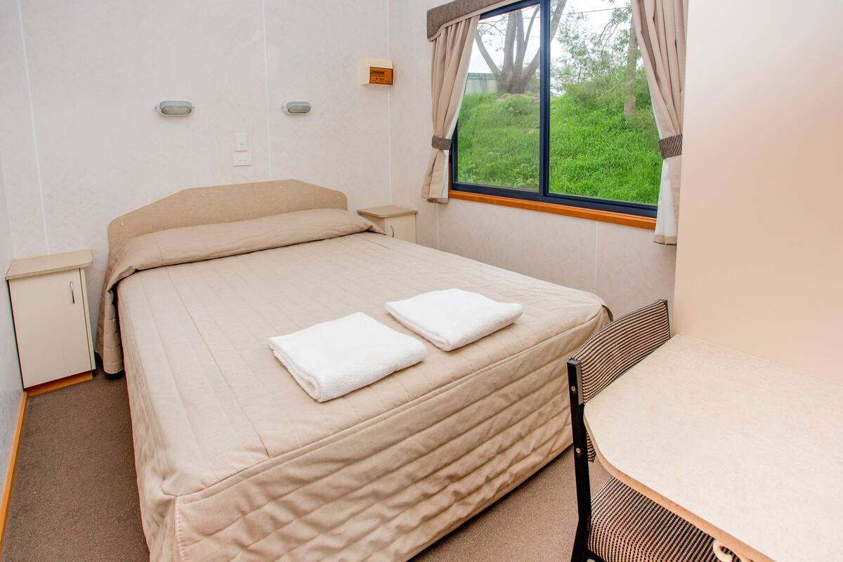 Un lit bien fait, avec deux oreillers et une serviette pliée, une petite table de chevet, une fenêtre avec des rideaux et une chaise au pied du lit.