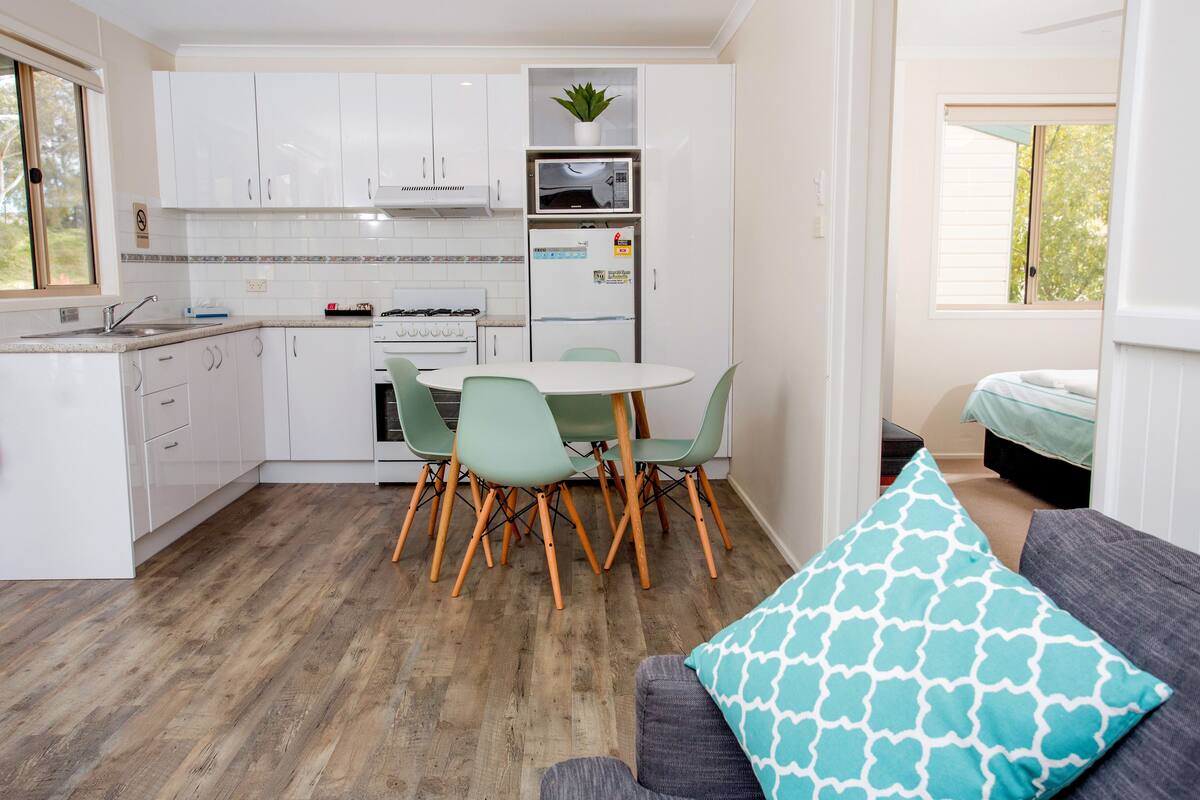 Une cuisine compacte avec des meubles blancs, un four à micro-ondes et un réfrigérateur. On y trouve une table à manger avec des chaises, ainsi qu ’un canapé gris agrémenté d’un coussin à motifs.