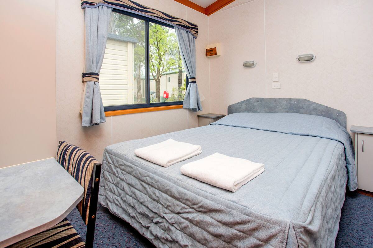 Un lit simple avec une couvre-lit grise et deux serviettes blanches pliées, une petite table, une chaise et une fenêtre avec des rideaux.