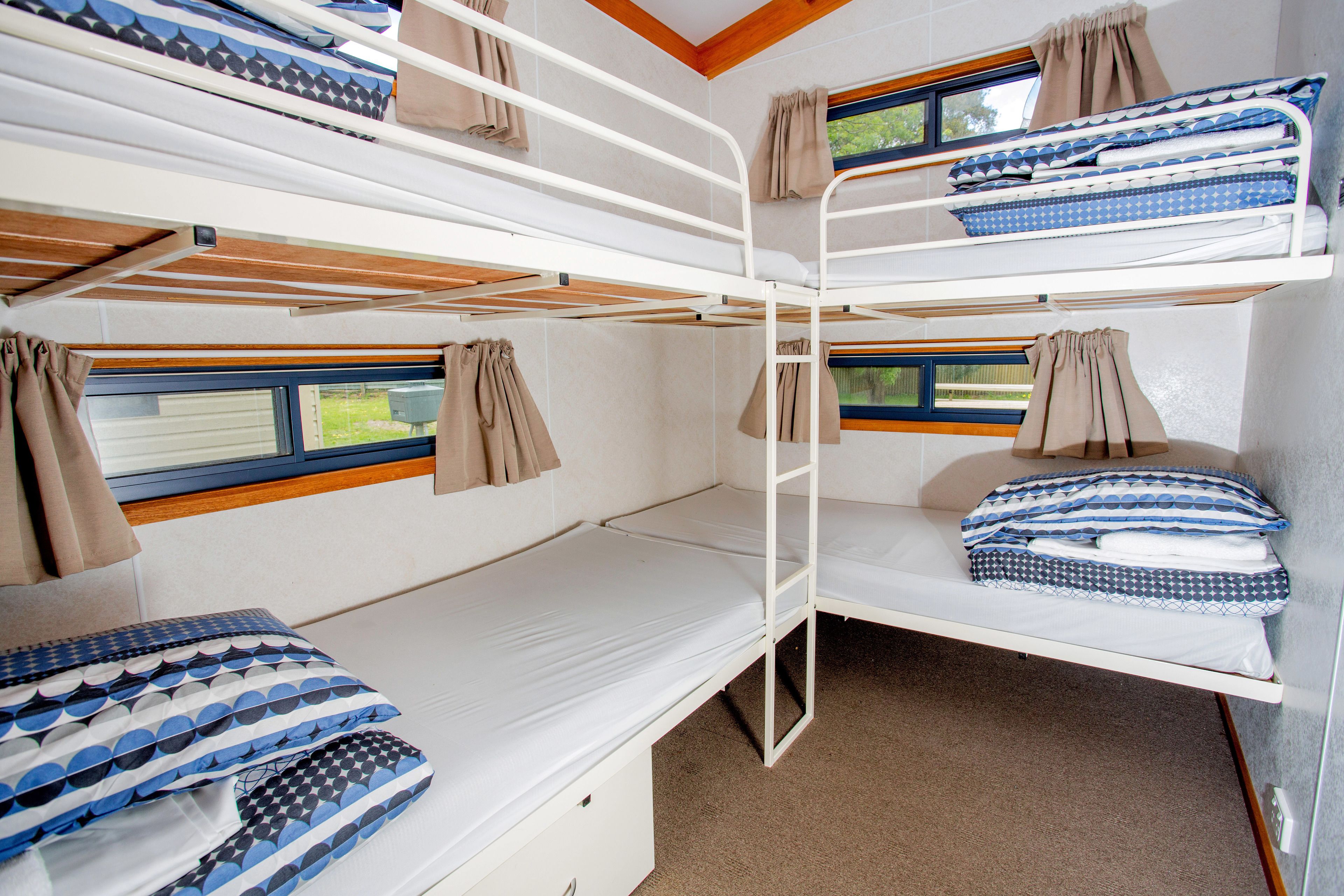 Een slaapkamer met een stapelbed, bestaande uit drie niveaus, elk met een matras en kussens. Er zijn ramen met gordijnen, en het plafond is van hout.