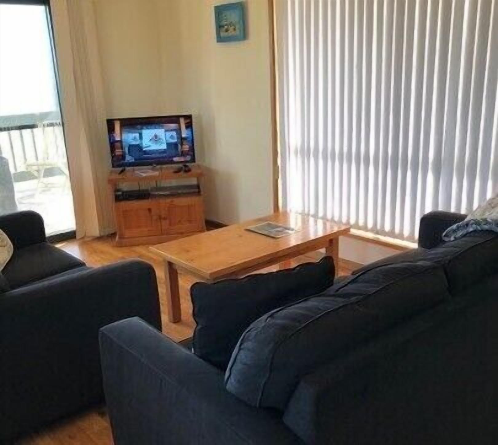 Una sala de estar con una mesa de centro de madera, un sofá de color oscuro, un televisor sobre un mueble de madera y una ventana con persianas verticales.