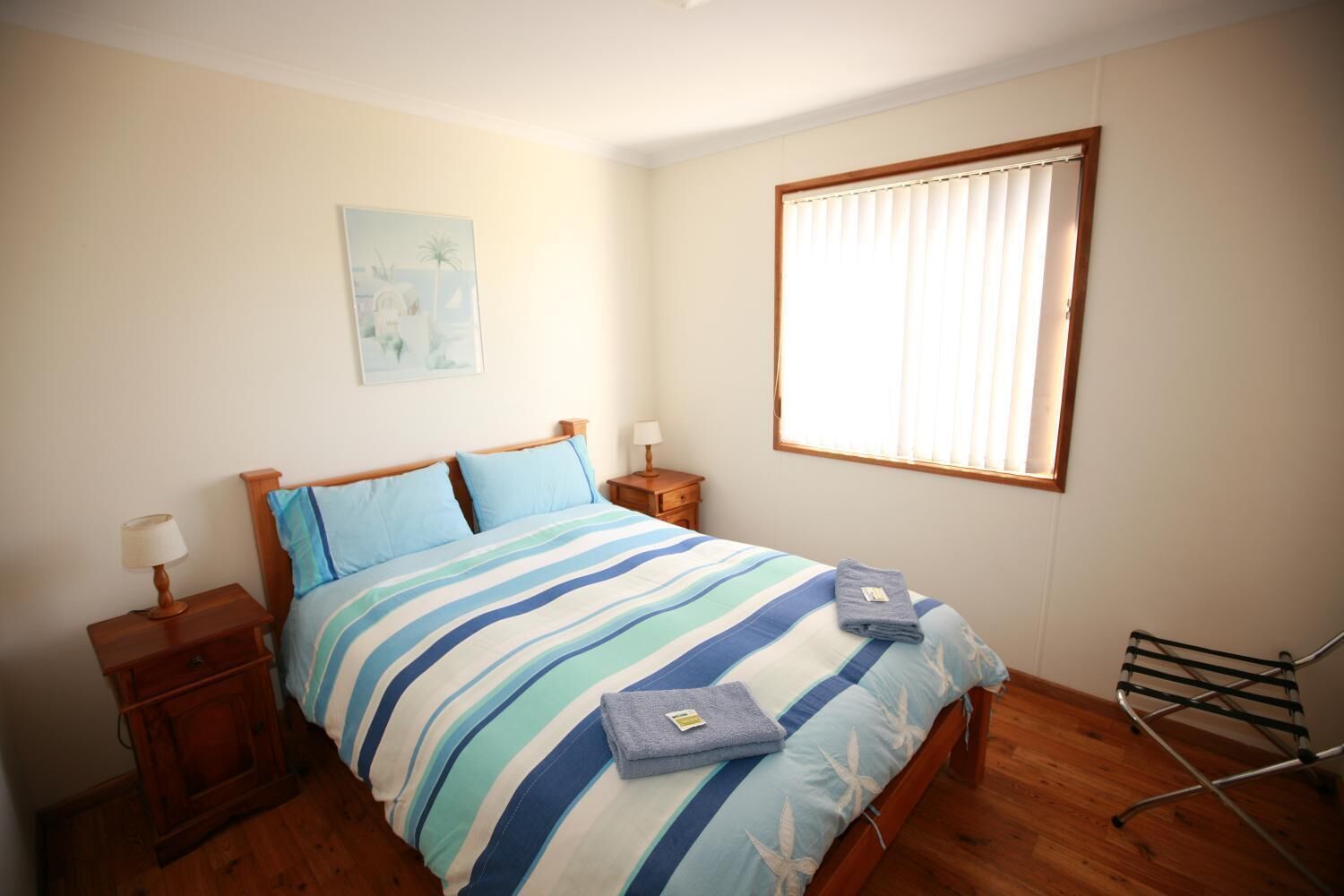 A bedroom with a bed, two bedside tables, a window with blinds, a wall-mounted artwork, and a wooden floor.
