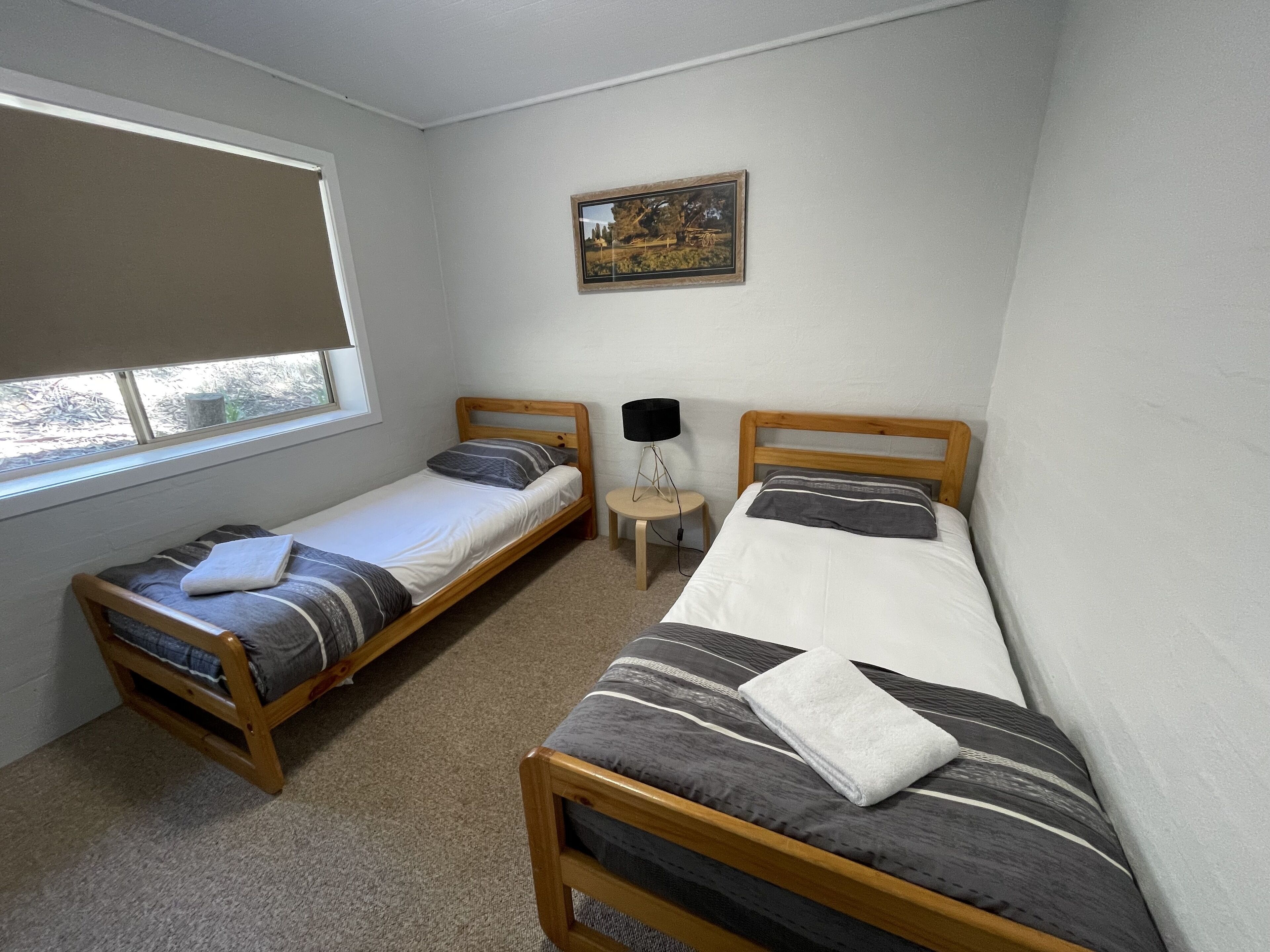A small, clean room with two single beds, a small table, a lamp, and a window.