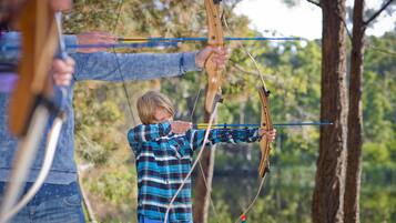 A person aiming a bow in an outdoor setting with trees in the background.