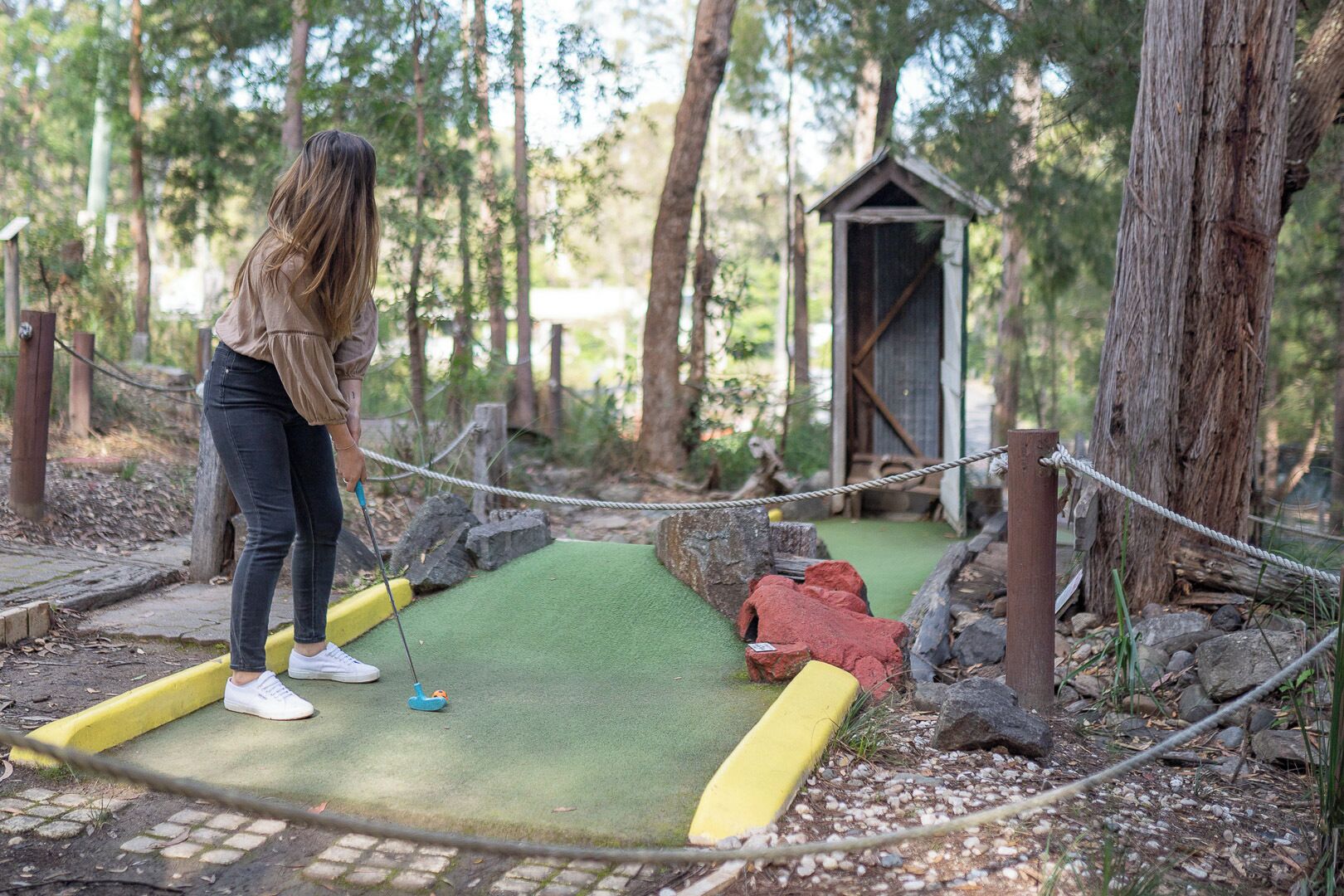 A person playing mini-golf in a wooded area with a small wooden structure in the background.