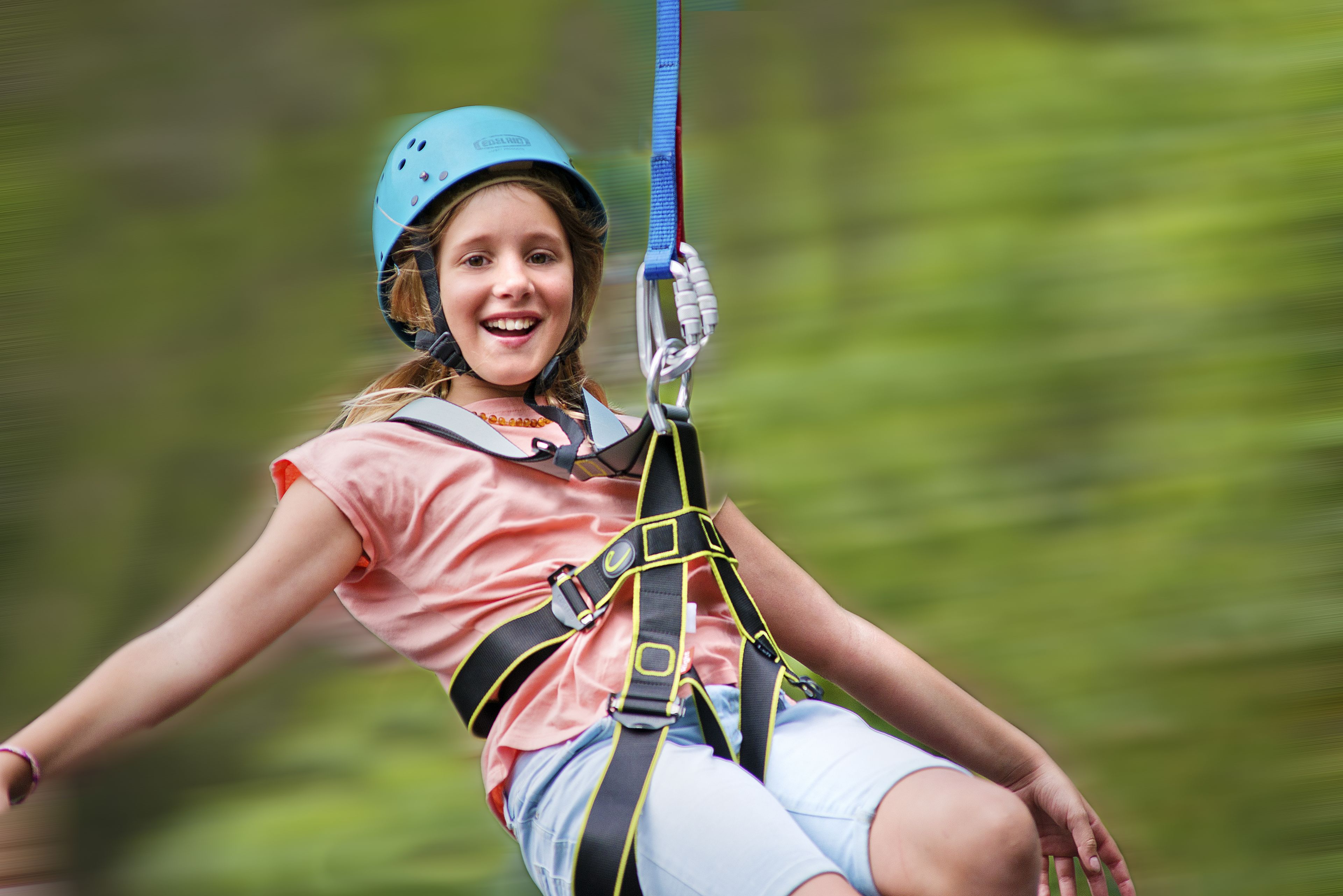 Eine Person mit Zipline-Ausrüstung und Helm, die ein pinkes Hemd und Jeansshorts trägt, vor unscharfem Grün im Hintergrund.