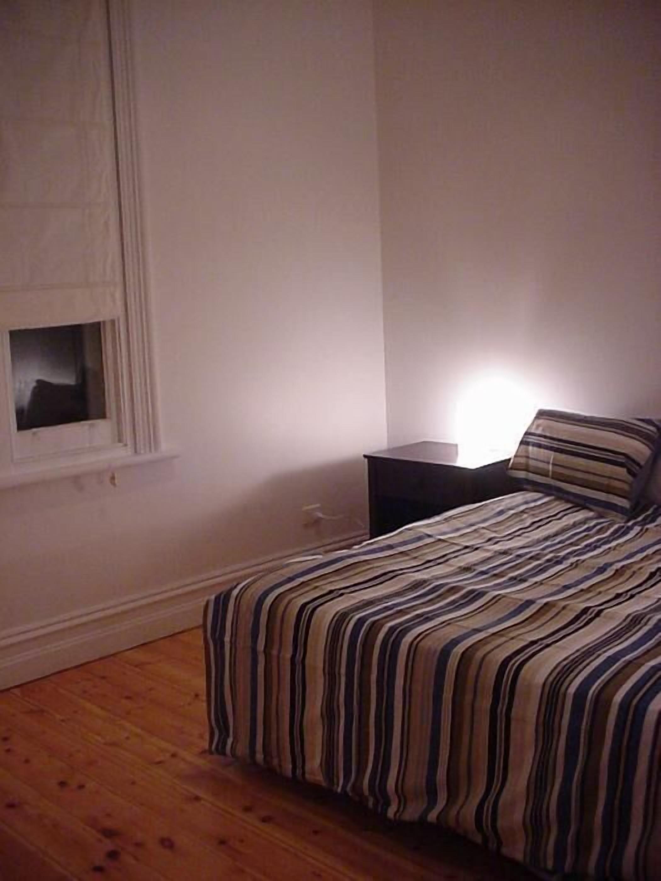A bedroom with a wooden floor, a bed with a striped bedspread, a nightstand, and a window with a view.