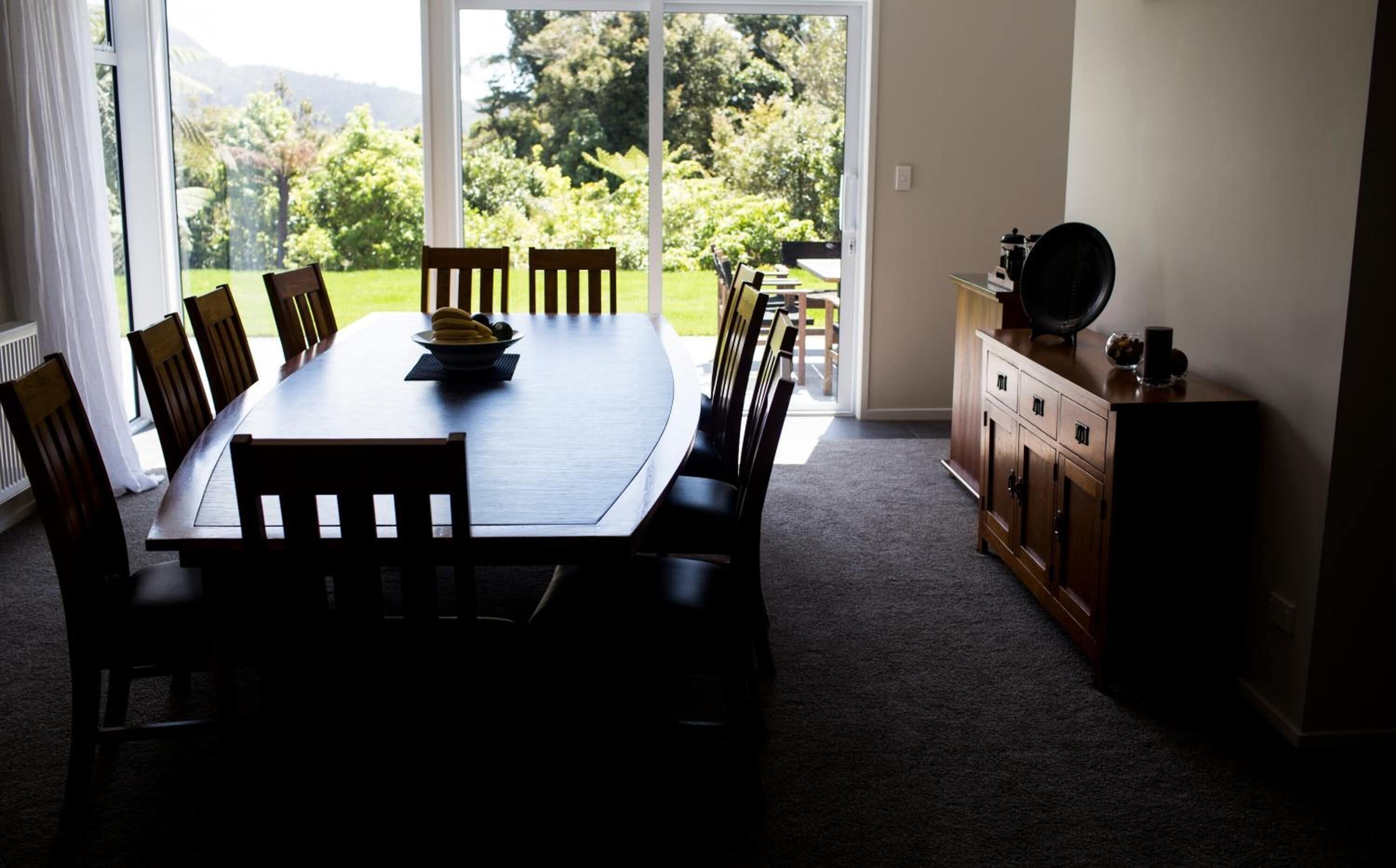 Uma área de refeições com uma mesa grande e cadeiras, um buffet com comida e vista para o verde através de grandes janelas.