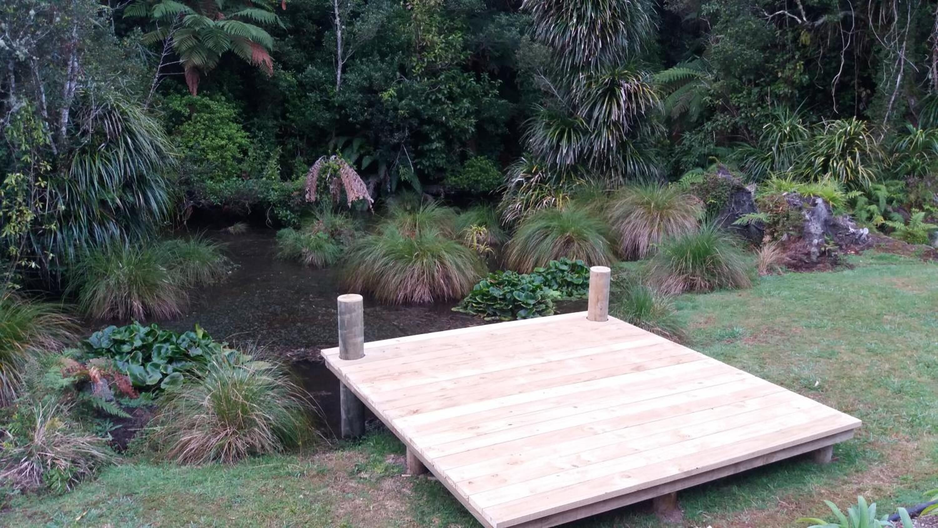 Une terrasse en bois, soutenue par deux piliers, entourée d’une végétation luxuriante et agrémentée d’un petit élément décoratif aquatique.