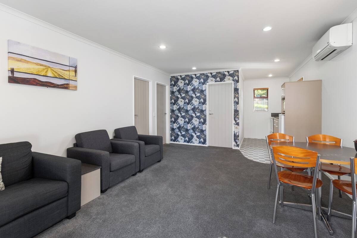 A modern hotel room with a grey carpet, a sofa, a dining table with chairs, and a kitchen area.