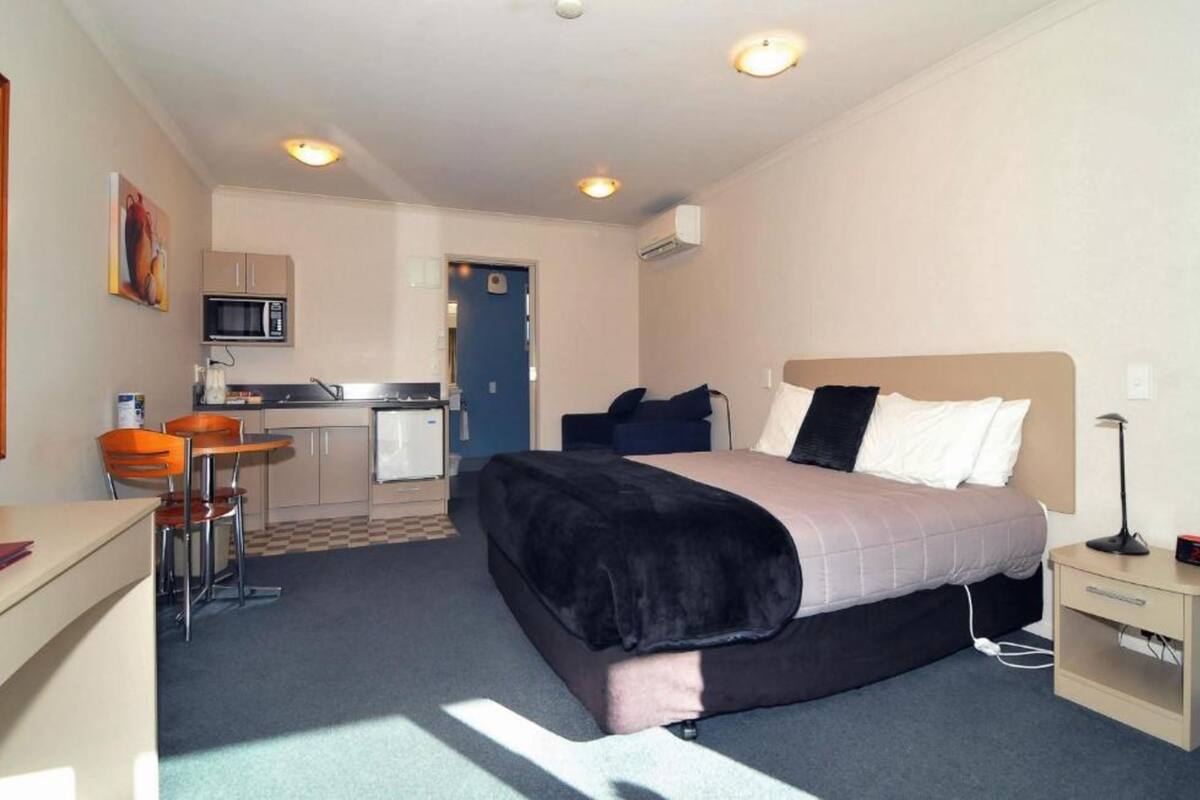 A hotel room with a bed, bedside table, desk, chair, kitchenette, and a door leading to a bathroom.