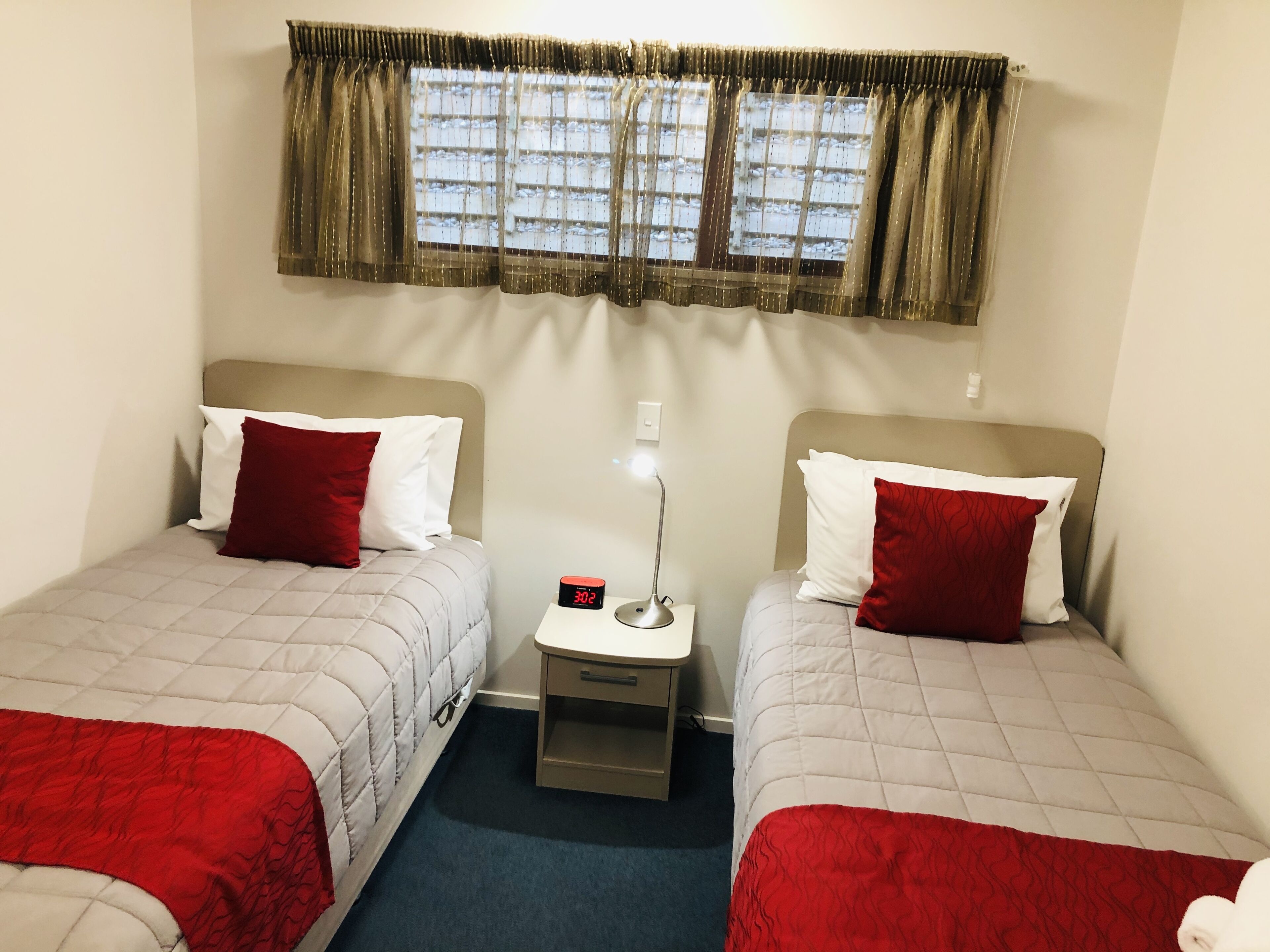 A twin room with two beds, each with a red and white pillow, a small bedside table with a lamp, and a window with a curtain.
