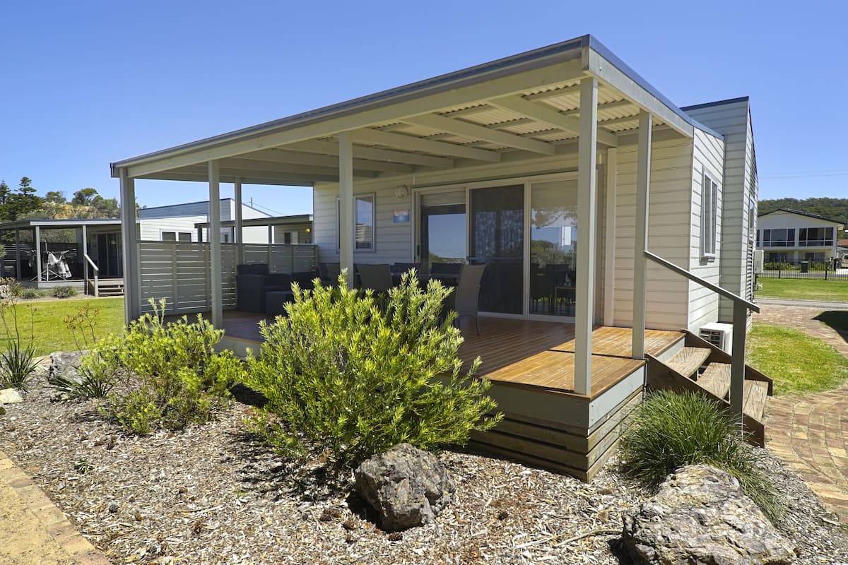 A modern house with a covered patio, a wooden deck, and a small garden with rocks and shrubs.