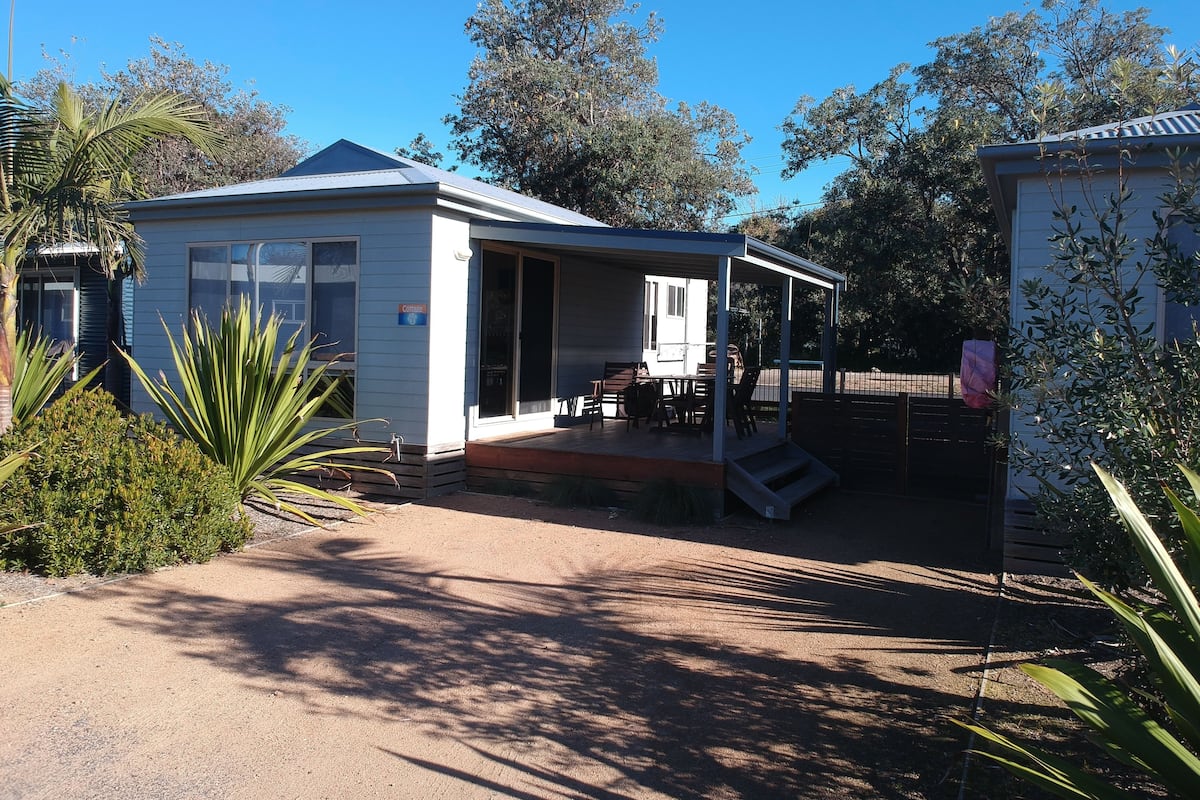A small, single-story building with a covered patio area, a fenced yard, and surrounding vegetation.