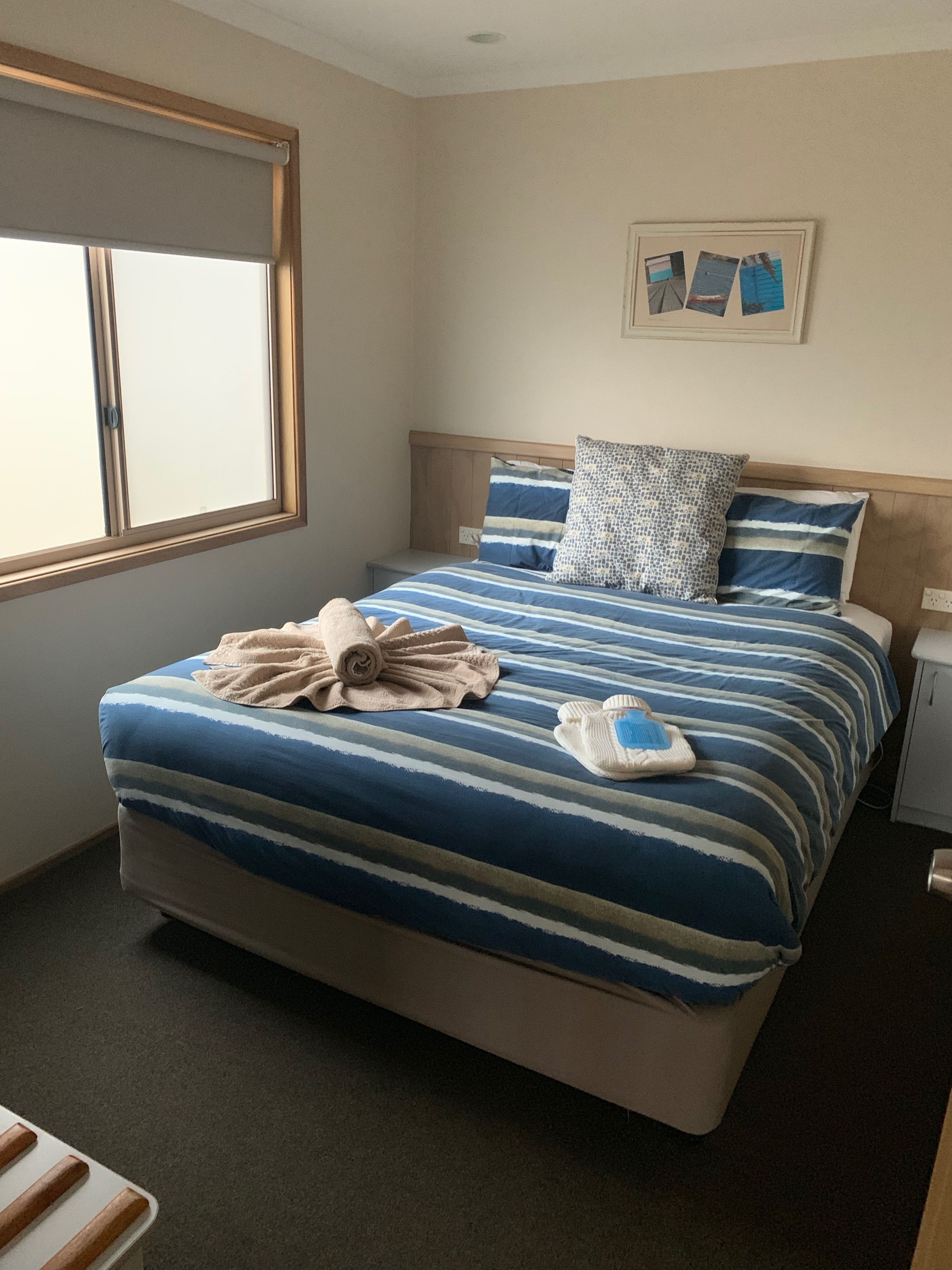 A neatly made bed with striped bedding and a rolled towel, a bedside table with a lamp, and a window with blinds.
