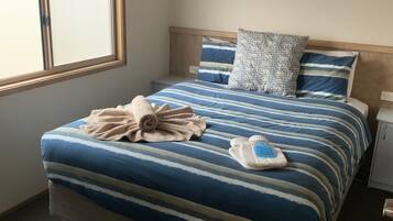 A neatly made bed with striped bedding and a rolled towel, a bedside table with a lamp, and a window with blinds.