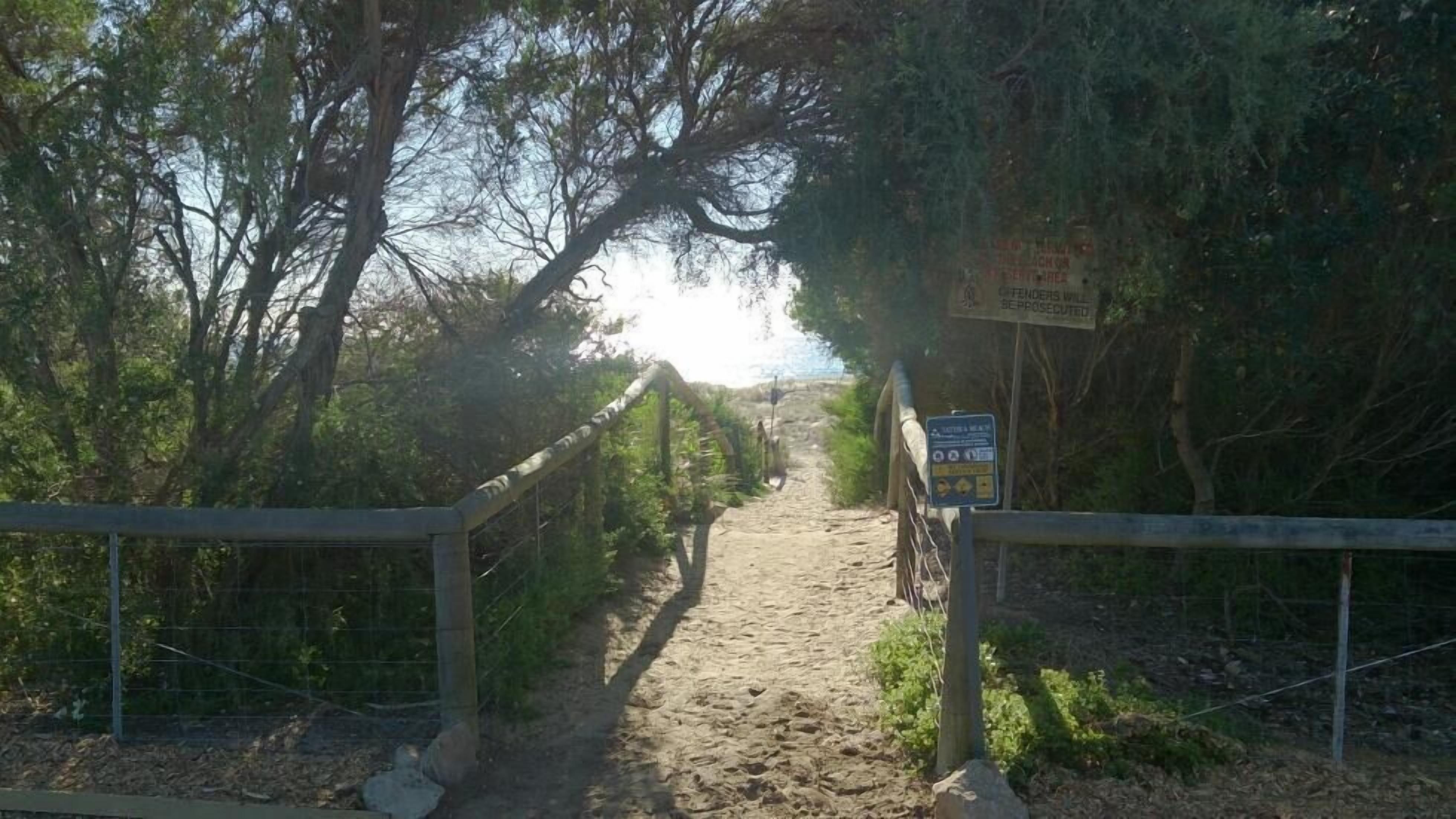 Uma trilha de areia serpenteia por uma área arborizada, com um portão de metal e uma placa.