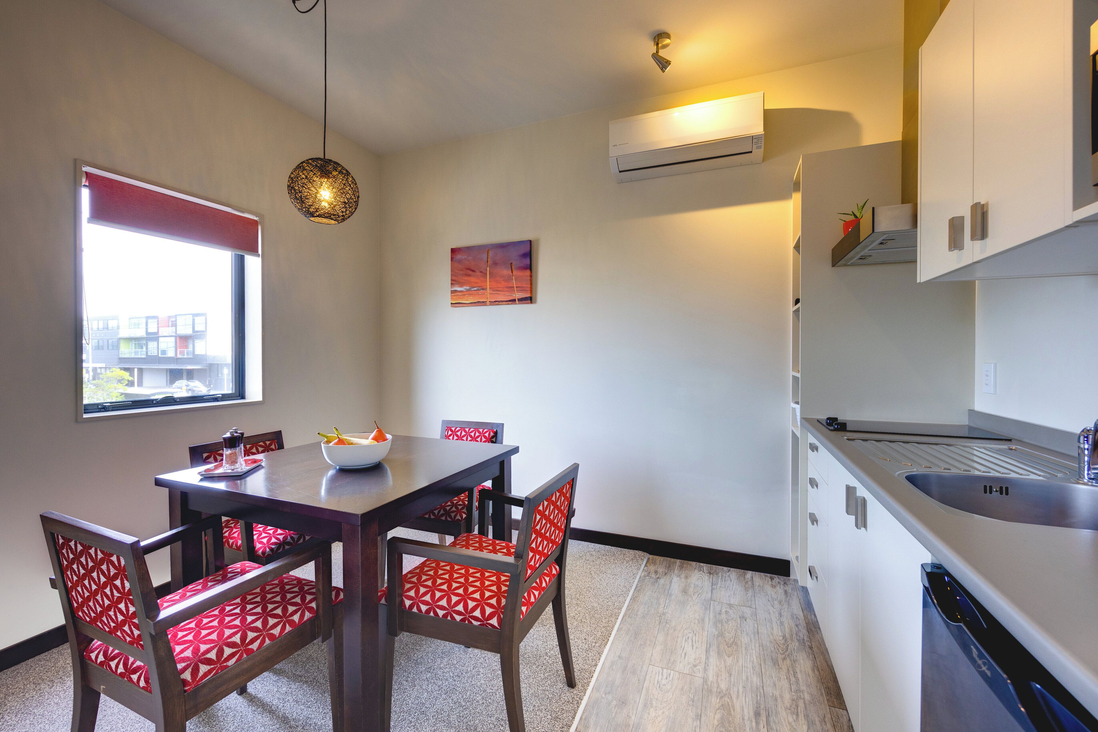 A compact kitchen with a dining area, featuring a modern design with white cabinets, a built-in dishwasher, and a dining table with red patterned chairs.