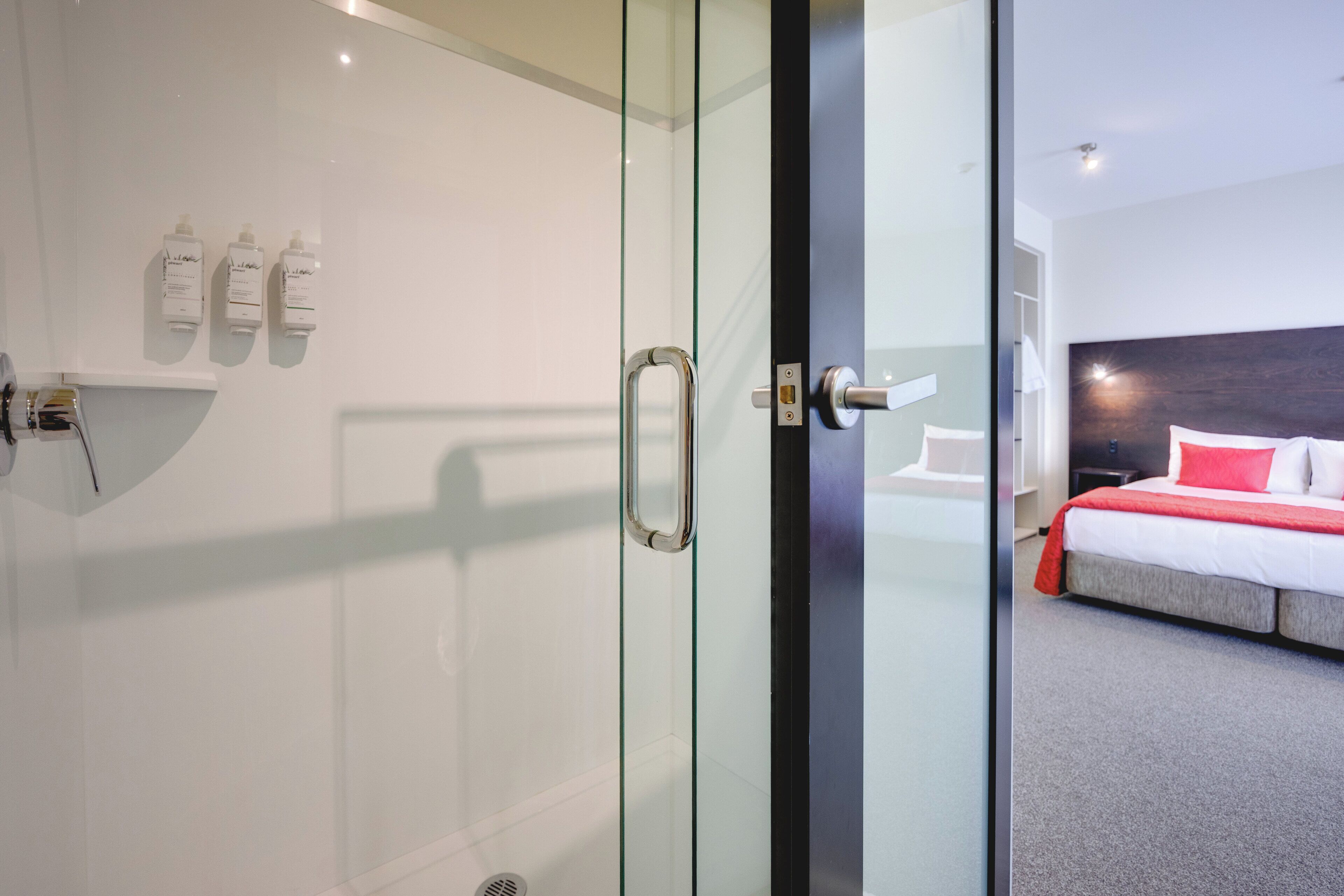 A modern hotel room with a glass shower enclosure, a bed with red and white bedding, and a white wall with three bottles of toiletries.