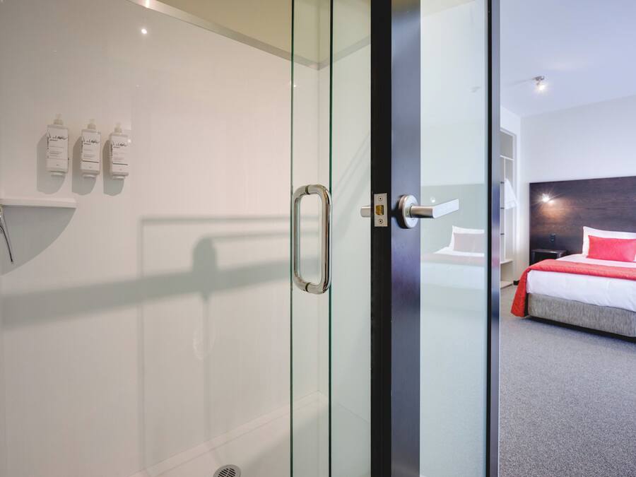 Una habitación de hotel moderna con cabina de ducha de vidrio, cama con ropa de cama roja y blanca, y una pared blanca con tres botellas de artículos de aseo.