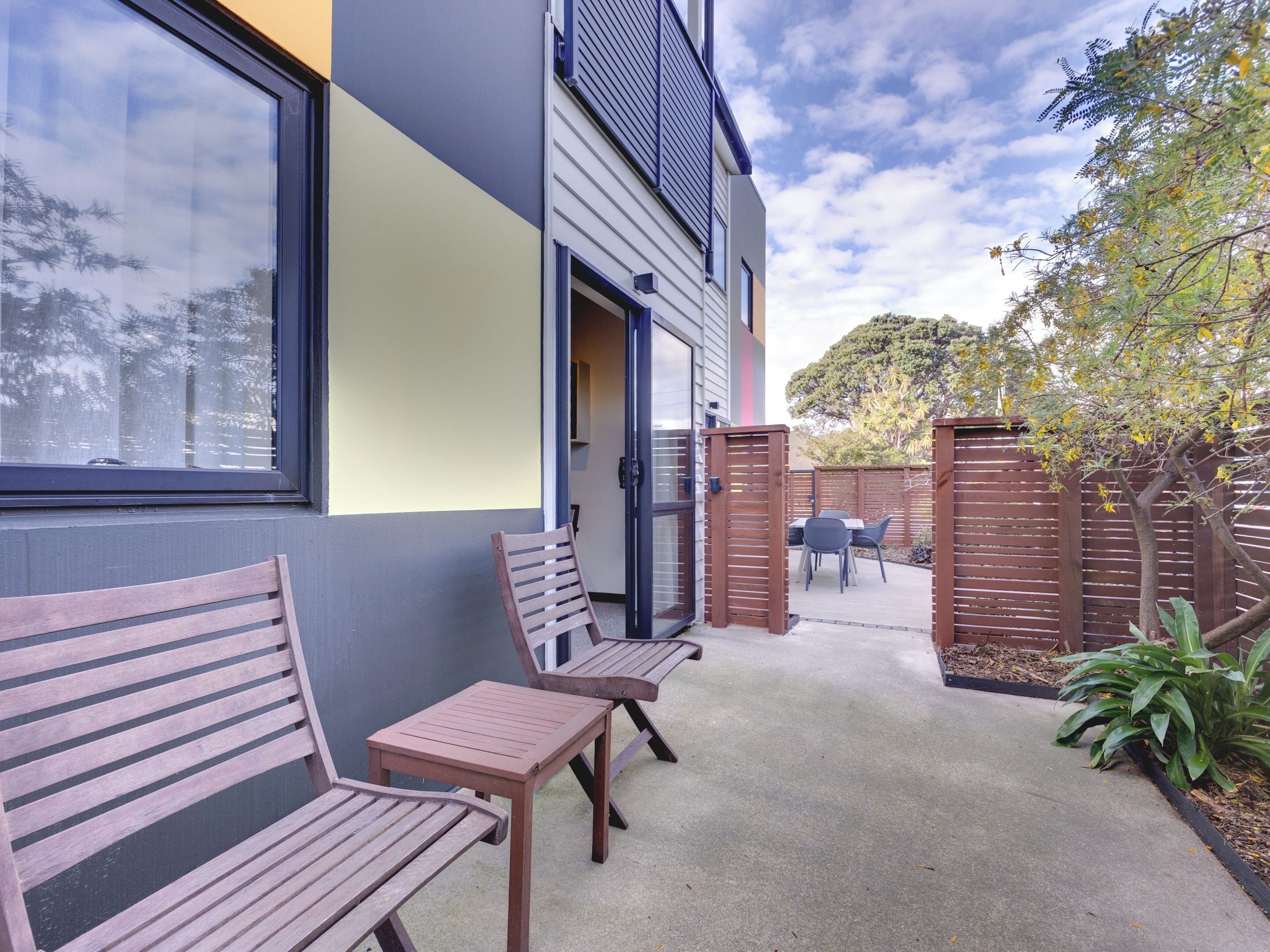 A patio area with wooden benches and a table, a sliding glass door, and a view of a tree and fence.