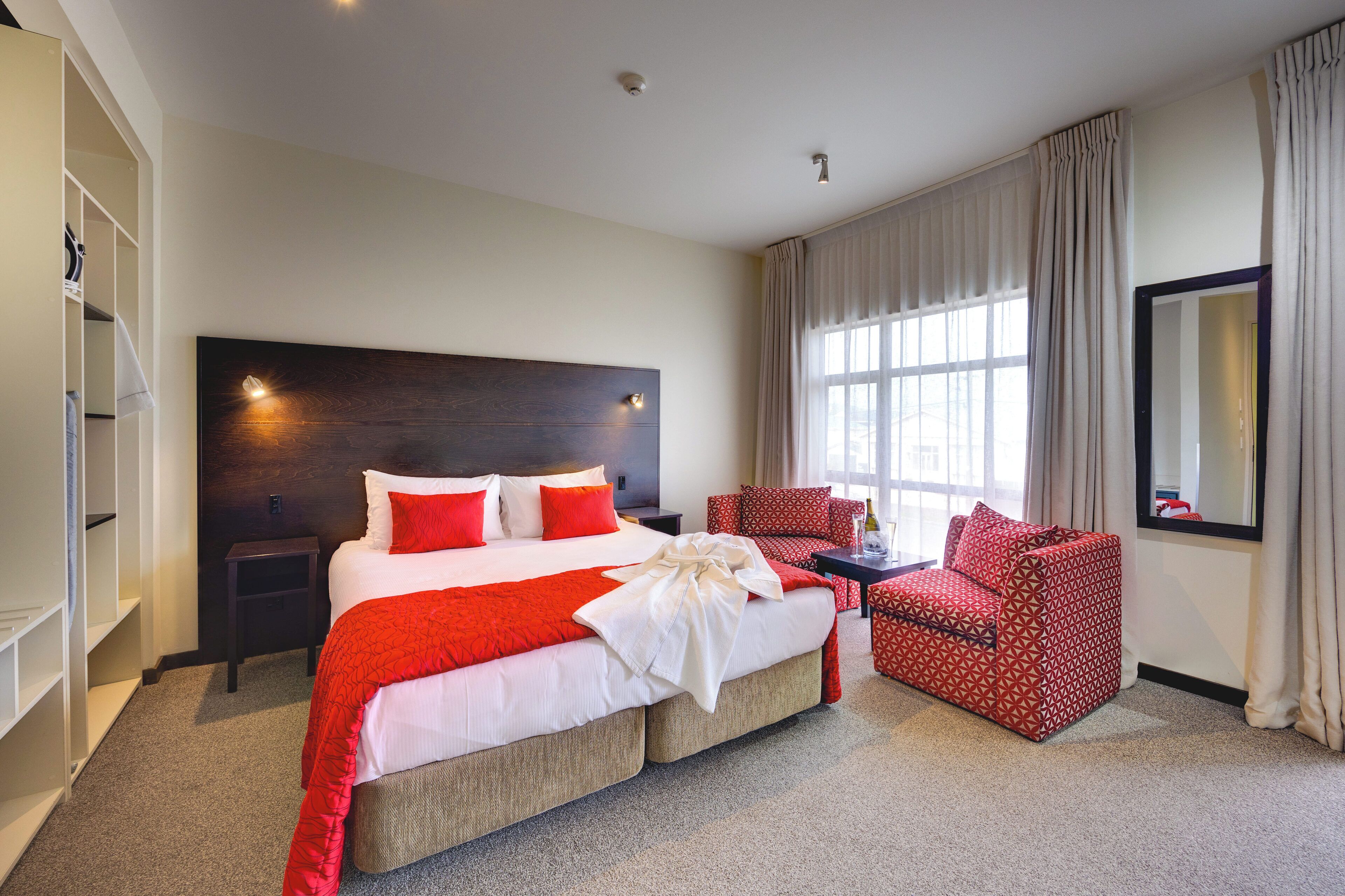 A hotel room with a large bed, a red armchair, a small table, and a large window with curtains.