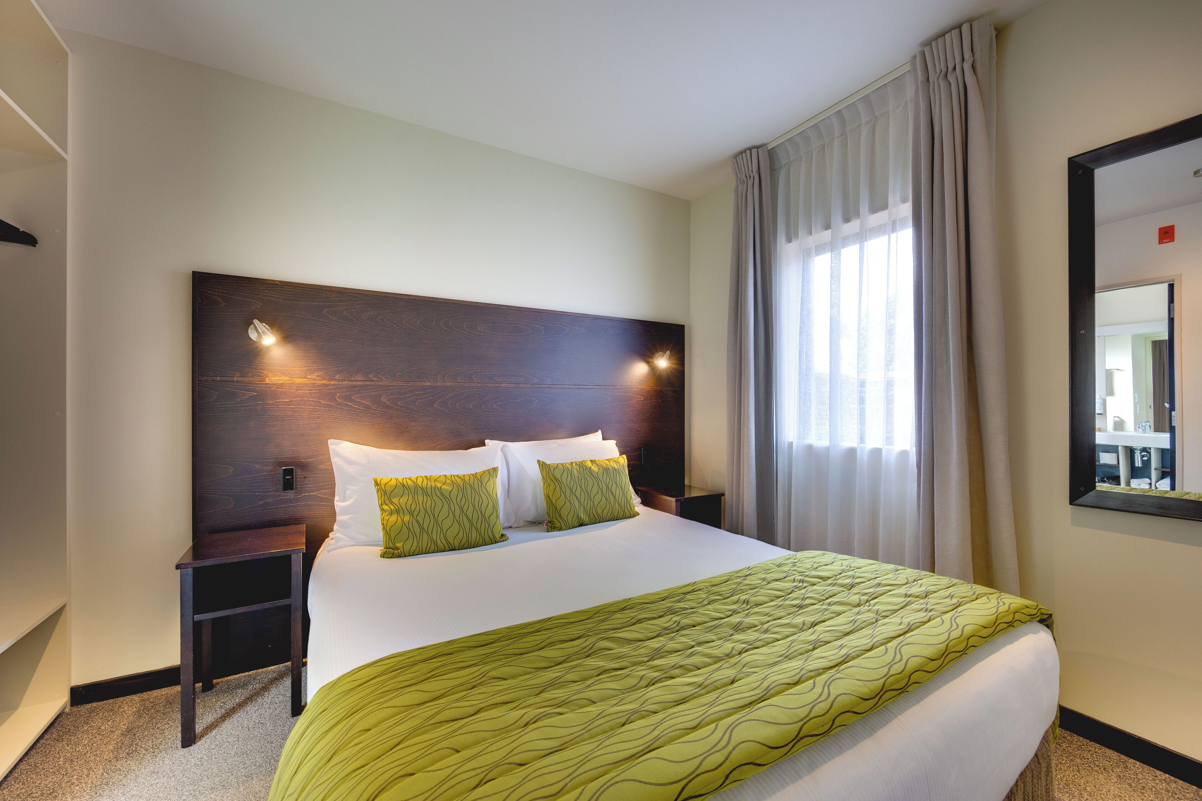 A hotel room with a large bed, two bedside tables, a headboard with wall-mounted lights, a window with curtains, and a mirror on the wall.