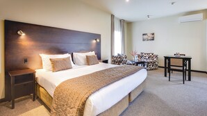 A hotel room with a large bed, a bedside table, a chair, a small table, and an air conditioning unit on the wall. - Ava Lodge (Lower Hutt)