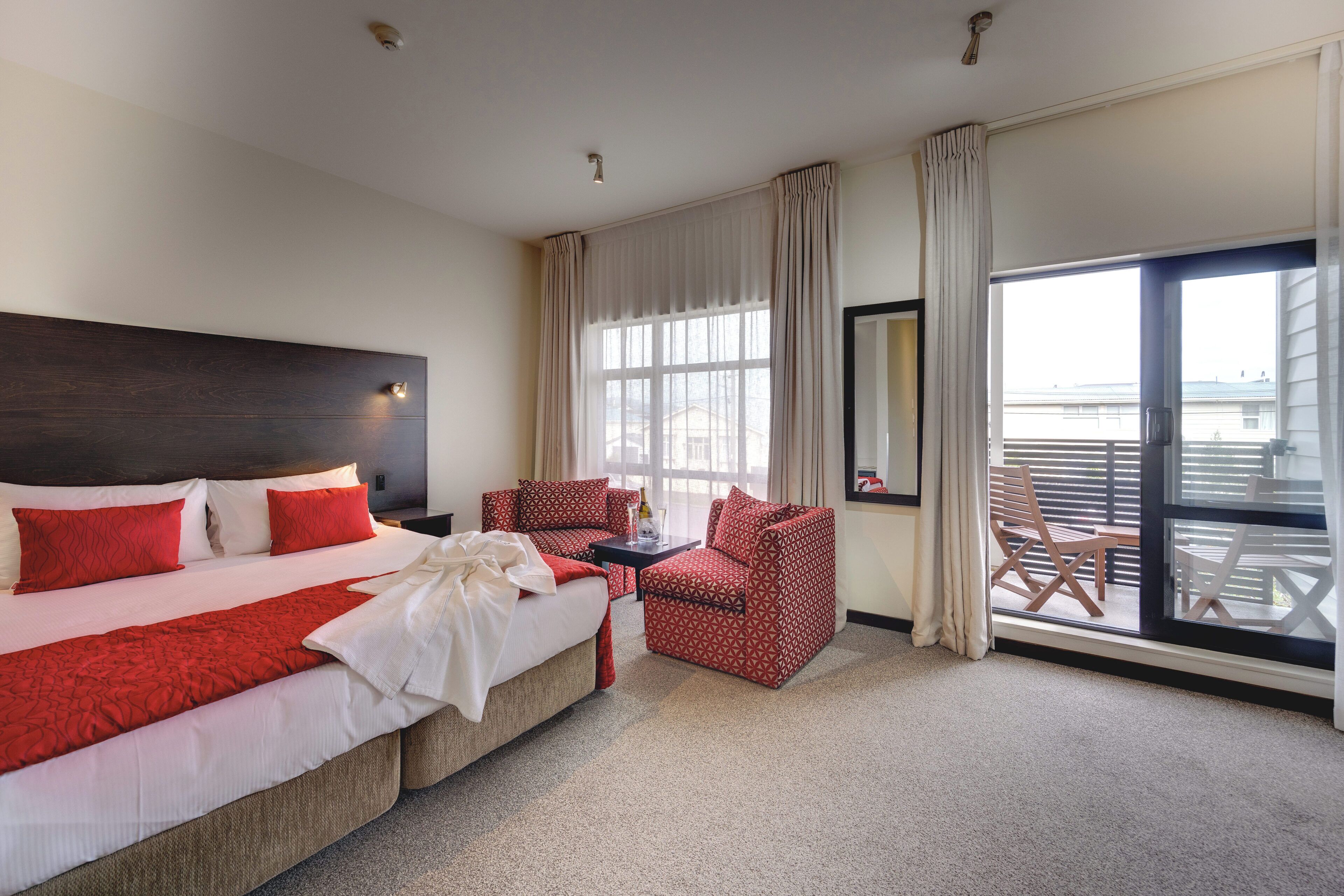 A hotel room with a large bed, red and white bedding, a small table, two red patterned chairs, and a balcony with wooden chairs.