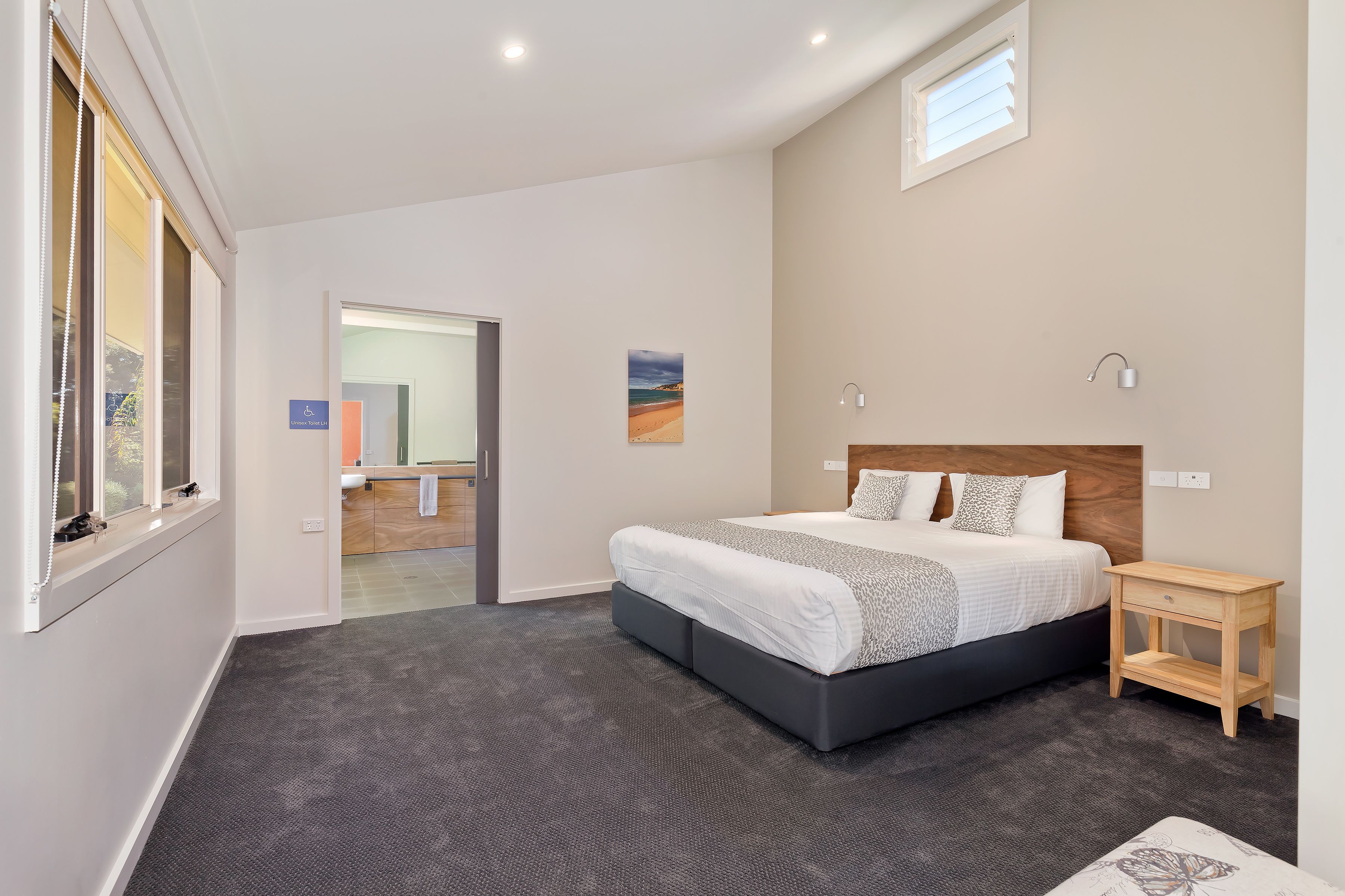 A modern bedroom with a large bed, a small bedside table, a window with blinds, and a painting on the wall.