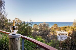 Varanda com uma garrafa de vinho e copos, com vista para uma paisagem costeira com árvores e um edifício.