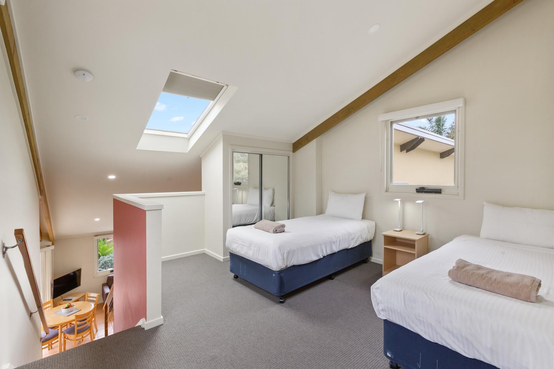 A bedroom with two beds, a skylight, a mirror, and a bedside table with a lamp.
