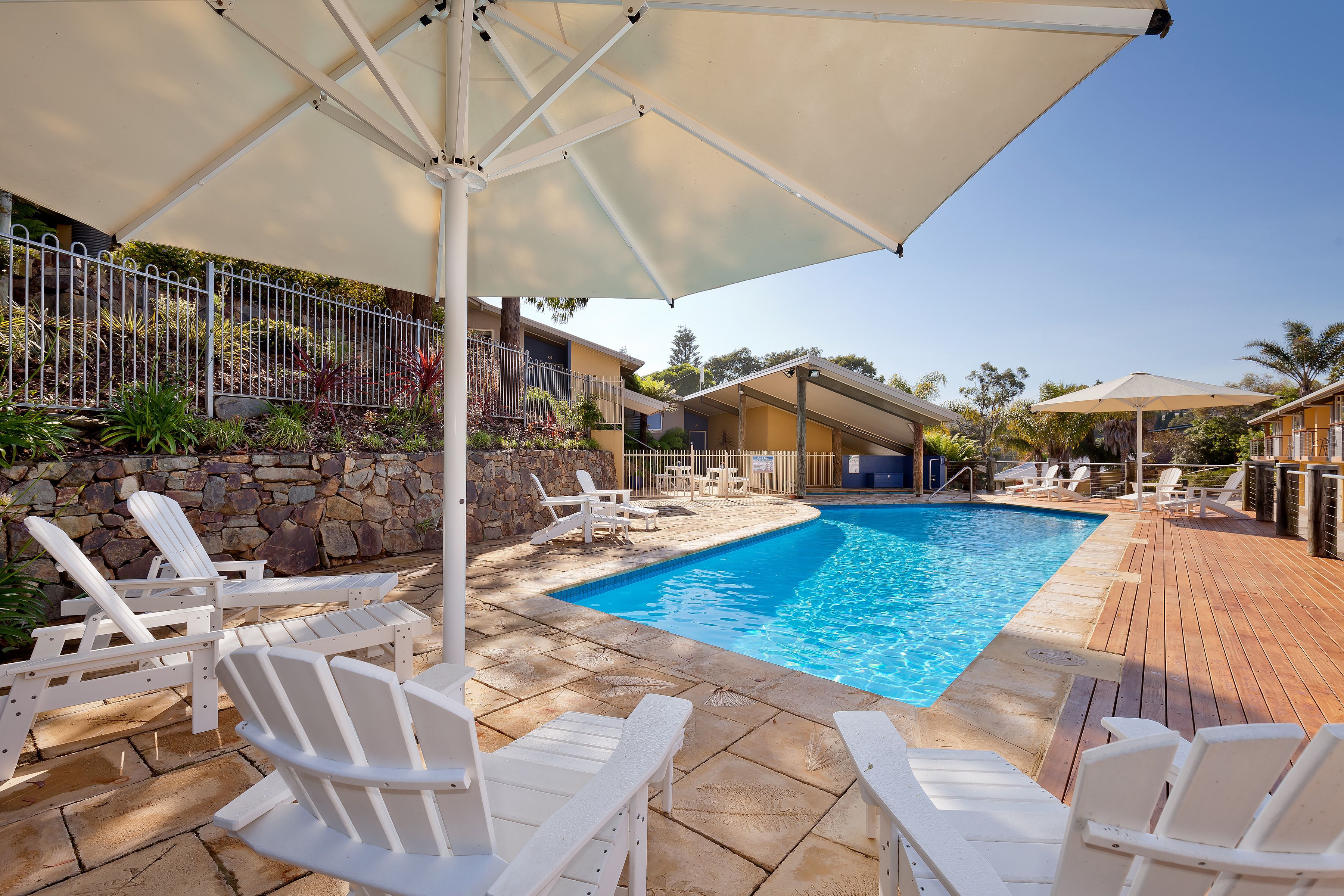 Zona junto a la piscina con sillas de descanso blancas, piscina y un porche cubierto con sombrillas.