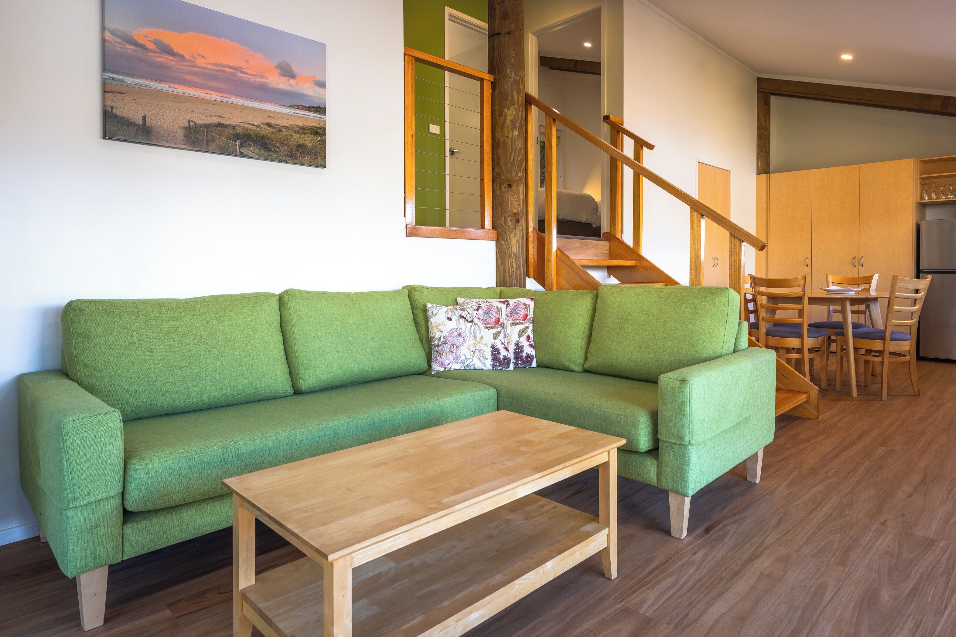A green sofa with a wooden coffee table, a dining area with wooden chairs, and a staircase with wooden railings.