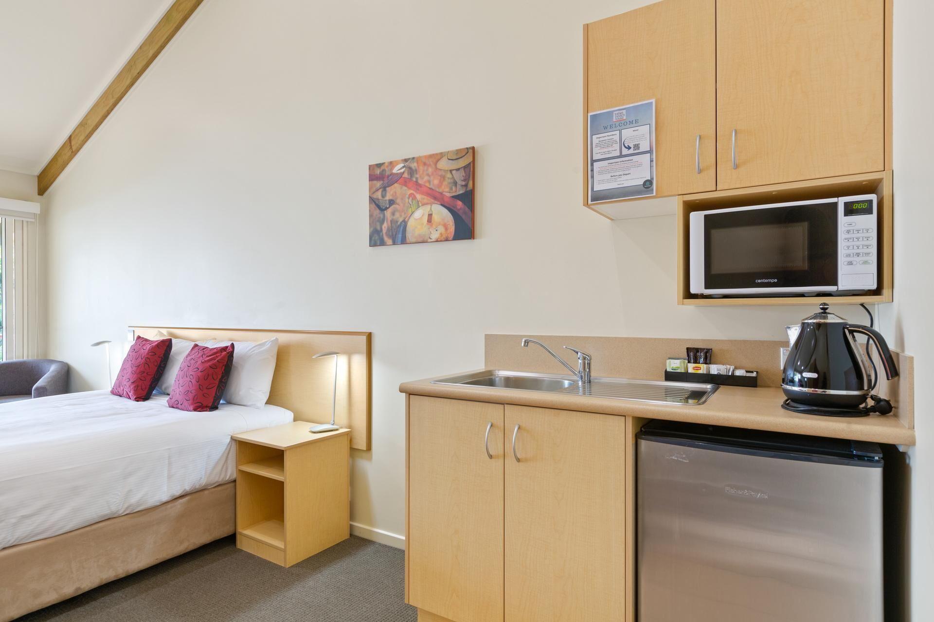Habitación de hotel compacta con cama, mesita de noche, pequeña kitchenette con microondas, lavamanos y refrigerador. Hay un cuadro en la pared y un cartel de bienvenida sobre la kitchenette.