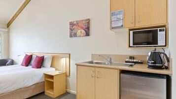 Habitación de hotel compacta con cama, mesita de noche, pequeña kitchenette con microondas, lavamanos y refrigerador. Hay un cuadro en la pared y un cartel de bienvenida sobre la kitchenette.