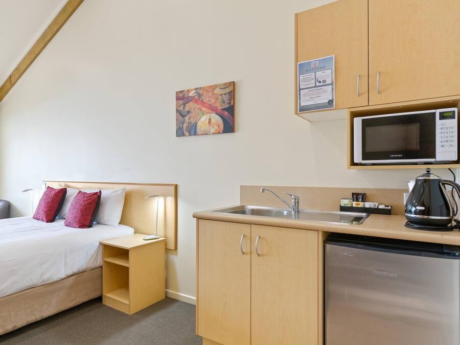 Habitación de hotel compacta con cama, mesita de noche, pequeña kitchenette con microondas, lavamanos y refrigerador. Hay un cuadro en la pared y un cartel de bienvenida sobre la kitchenette.