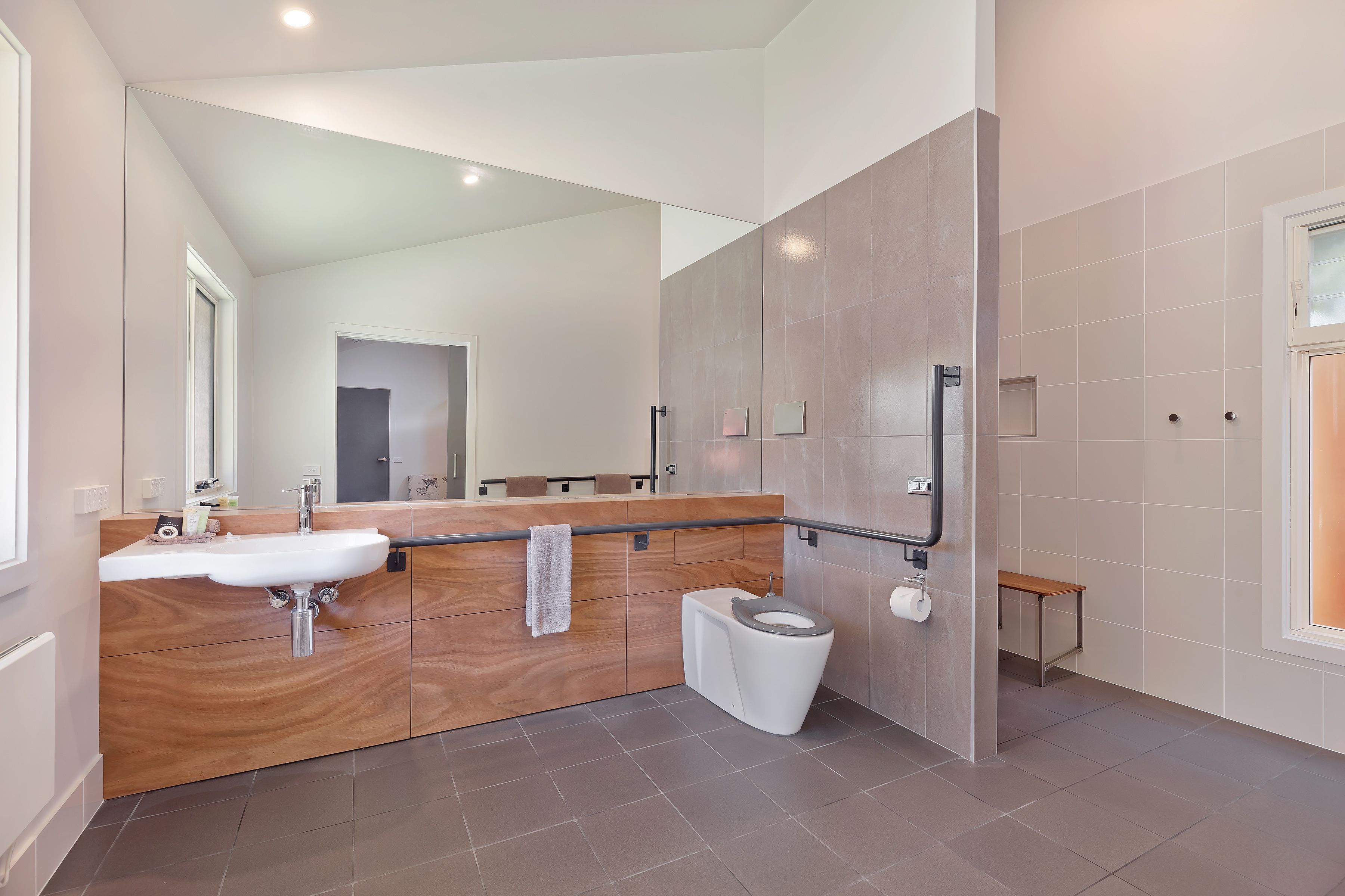 A modern bathroom with a wooden vanity, a white sink, a toilet, and a small bench.
