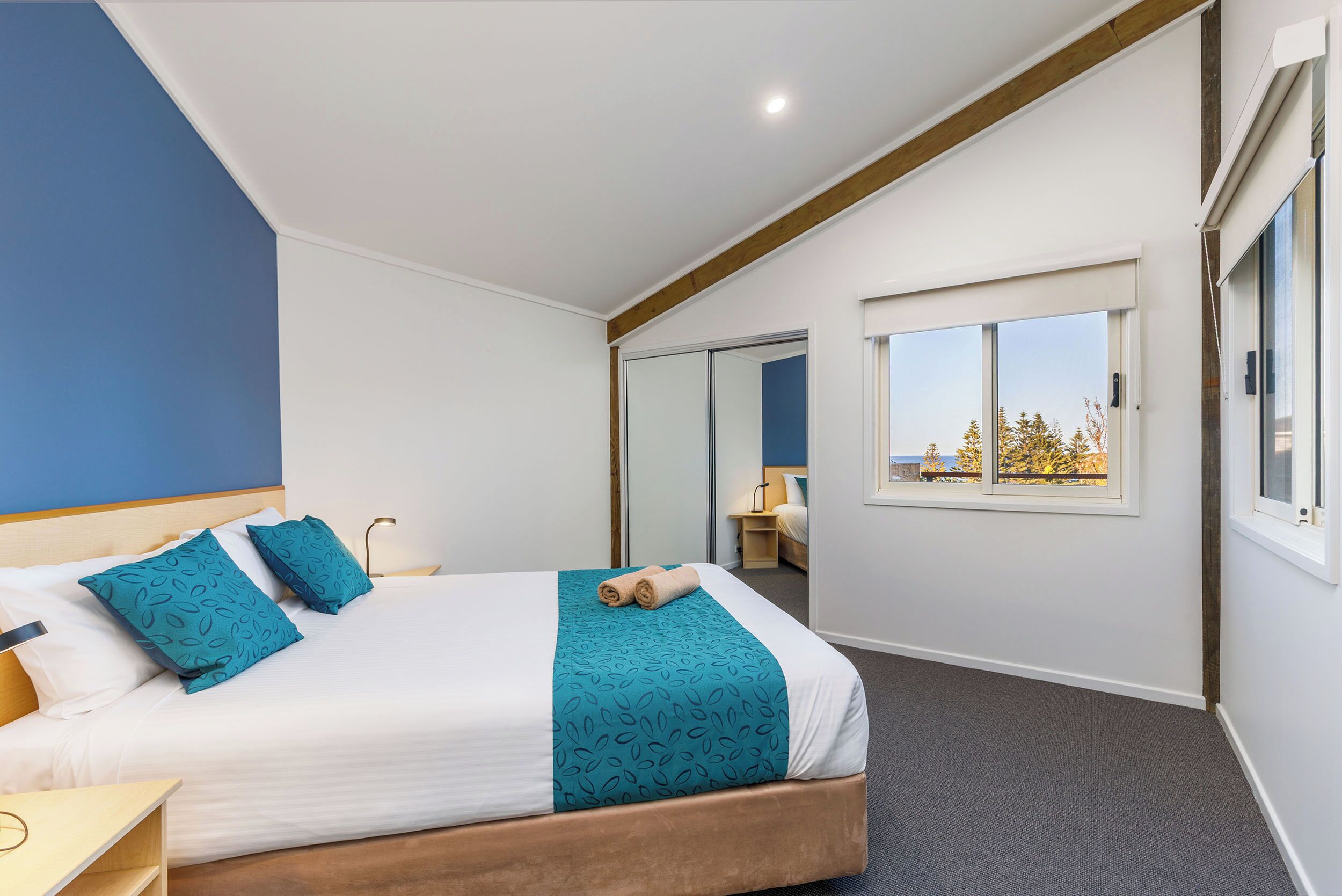 A modern hotel room with a large bed, a view of the outdoors, and a wooden ceiling beam.