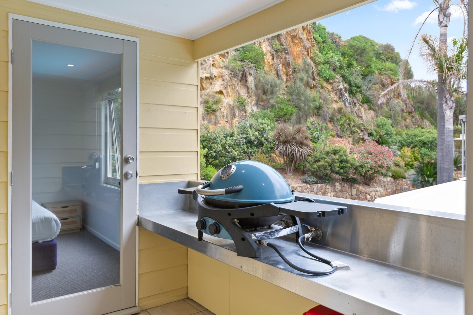 Ein Balkon mit Grill, eine Tür zu einem Raum und Blick auf einen felsigen Hügel mit Vegetation.