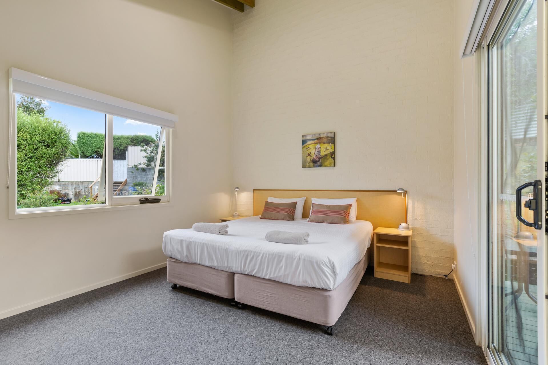 A bedroom with a large bed, a bedside table with a lamp, a window with a view of greenery, and a painting on the wall.