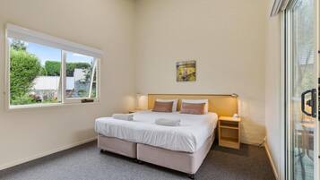 A bedroom with a large bed, a bedside table with a lamp, a window with a view of greenery, and a painting on the wall.