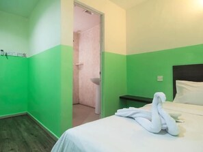 Une chambre d’hôtel aux couleurs vert et blanc, avec un lit recouvert de draps blancs, une table de chevet et une serviette posée sur le lit.