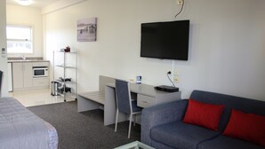 A compact hotel room with a kitchenette, a flat-screen TV, a grey sofa with red cushions, and a glass coffee table. - Hawera Central Motor Lodge (Hawera)