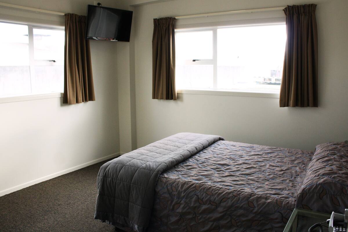 Ein Hotelzimmer mit einem Bett, einem an der Wand befestigten Fernseher und zwei mit Vorhängen versehenen Fenstern.